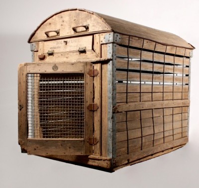 An unusual antique wooden dog carrier, by A. Backus, Jr., &amp; Sons, dating from the early 1900’s and featuring a signed medallion on the front identifying the maker. Absalom Backus, Jr. (1824-1901) and his sons were manufacturers of dog crates and woven boxes in Detroit, MI. Henry N. Backus patented this animal crate in 1902, patent number 698782 – designed for “shipping fine dogs and other valuable animals.” This dog kennel features an arched roof, with a door in the front and a spring-loaded chute above for food (labeled “Dog Biscuit, Water Here, Pull Open to Feed”). The edges of the crate are reinforced with metal; however, this apparently didn’t deter a feisty previous occupant, who appears to have chewed through the crate at the edge of the door, and also managed to remove a few boards from the floor of the cage. This dog cage measures 28 ¼” wide, 40 ½” long, and 36” tall.